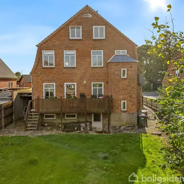 En murstensbygning med flere etager, står i en have med grøn græsplæne. Bygningen har en bagterrasse med træmøbler, og omkring er der andre huse og træer.