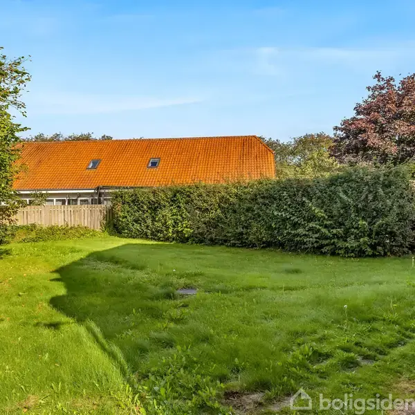 Græsplæne strækker sig over et indhegnet område, omkranset af grønne hække og træer. I baggrunden ses et hus med rødt tag under en klar blå himmel.
