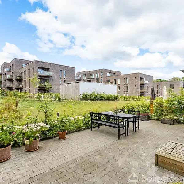 Terrassemøbler står på flisebelagt terrasse, omkranset af blomsterpotter og grønne buske, med udsigt til moderne lejlighedsbygninger af mursten. Himmel med hvide skyer ses i baggrunden.