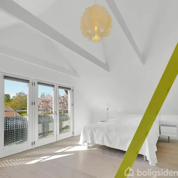 Et soveværelse med en hvid seng står stille under et skråt loft og lyse bjælker. En gul stige fører opad, mens store vinduer viser udsigt til en have med træer.