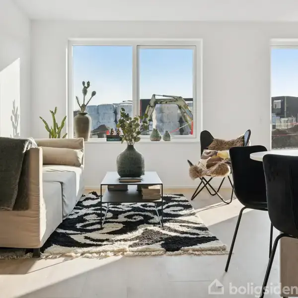 Stue med beige sofa og glasbord, omgivet af grønne planter og dekorationer, belyst af naturligt lys fra store vinduer med udsigt til omkringliggende bygninger. Sorte stole ved et spisebord.