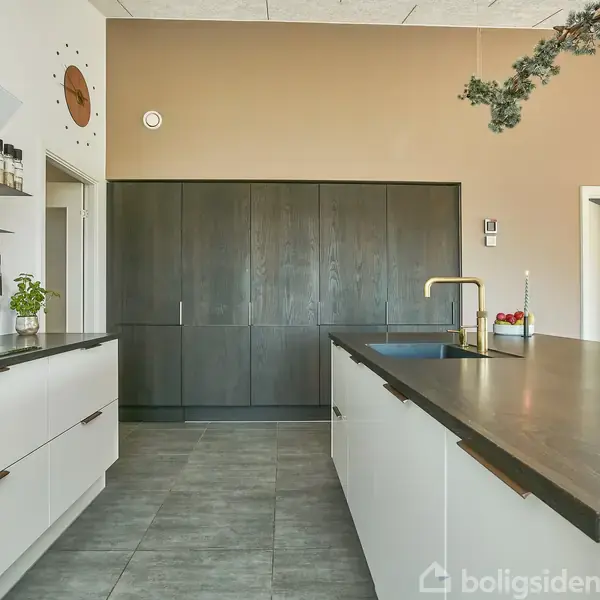 Langt spisebord med moderne stole står i et lyst køkken-alrum. På væggen hænger et verdenskort og to store pendellamper. Blomster pryder bordet.