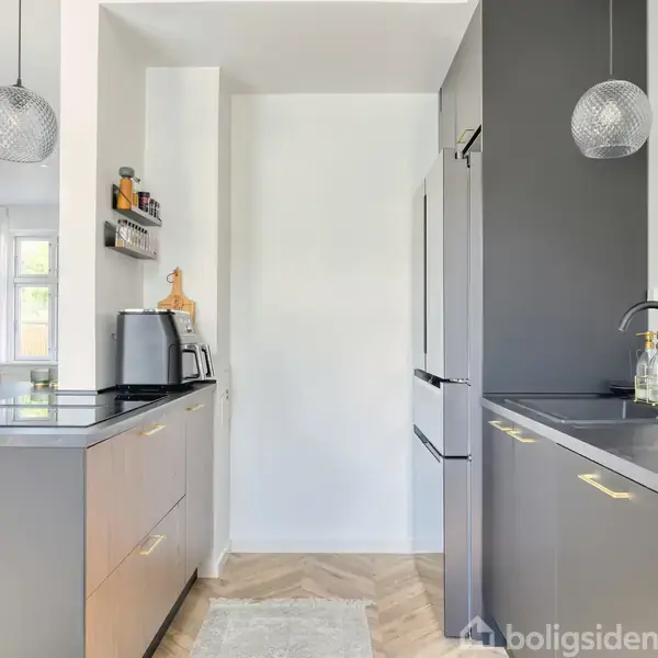 Køkken med lysebrune skabe og sorte bordplader, monteret med to glaspendler. En ovn og mikrobølgeovn står til venstre; køleskab til højre. Rummelig og moderne indretning.