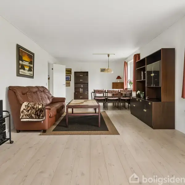 En stue med en brun lædersofa med et tæppe, sofabord og reol. Spisebord med stole i baggrunden; lysindfald fra vinduet med røde gardiner. Ingen tekst.