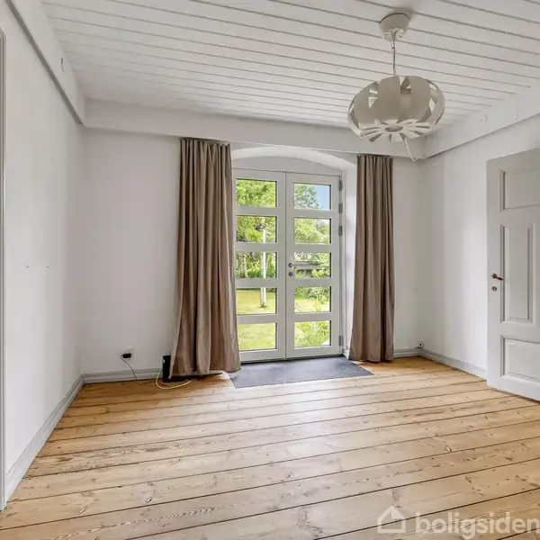 Et rummeligt, lyst værelse med trægulv og hvidt loft. En buet glasdør åbner til en grøn have. To døre fører til andre rum, og brune gardiner hænger ved døråbningen.