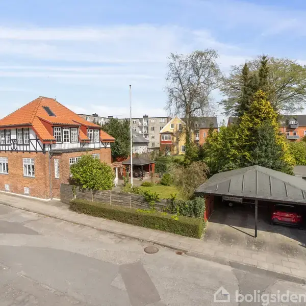 Et to-etagers murstenshus med rødt tegltag står på en villavej. En carport med en bil er til højre, omgivet af træer og have.