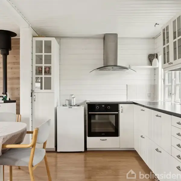 Køkken med hvide skabe og grå bordplade, integreret komfur og emhætte; spisebord med stole i forgrunden; pilleovn til venstre ved siden af en vitrineskab; lyst træ- og hvid vægpanel.