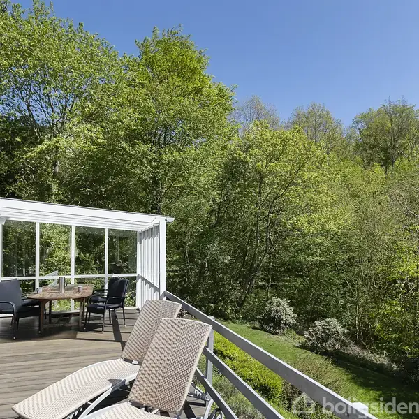 Terrasse med liggestole og et bord med stole. Handling foregår på en træterrasse omgivet af frodige grønne træer og blå himmel, hvilket skaber en afslappende atmosfære.