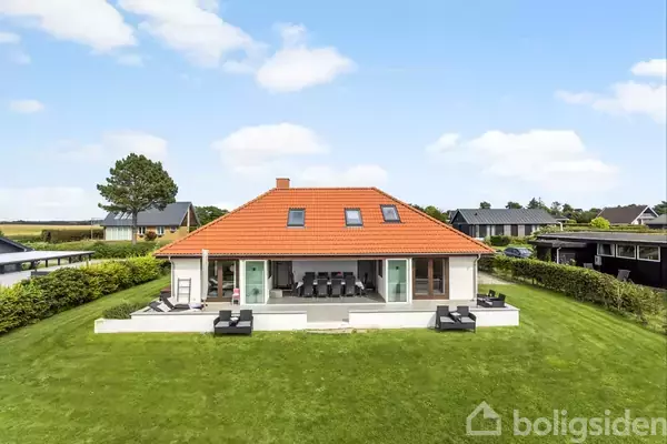 Et moderne hus med rødt tag står på en velplejet græsplæne. Store vinduer og terrassemøbler pryder den forreste del. Baggrund viser et boligområde med hække og træer.