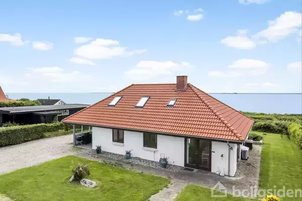Et hvidt hus med rødt tegltag ligger stille i en have med græsplæne. I baggrunden ses havet og en blå himmel med skyer, omgivet af grønne buske.