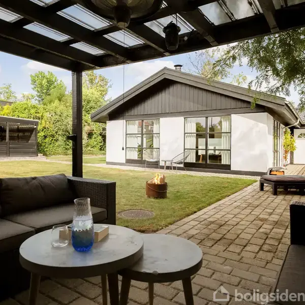 Sofa står på en overdækket terrasse; foran står et bord med en karaffel og bog. I baggrunden ses et moderne hus, plæne og liggestol i en have.