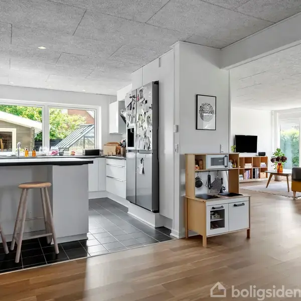 Køkken med to barstole ved en køkkenø, stort køleskab dekoreret med magneter, i sammenhæng med et åbent opholdsområde ved siden af, der har store vinduer og en gul sofa.