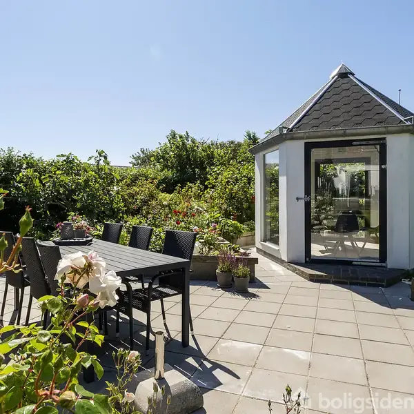 Et havepavillon står stille omgivet af grønne buske. Ved siden af er et bord med seks stole på en flisebelagt terrasse under en klar blå himmel.