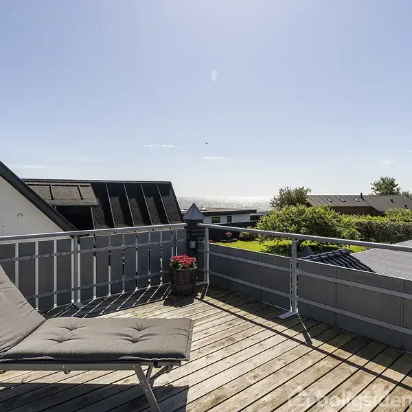 En liggestol står på en træterrasse med udsigt over hustage og grønne områder i solskinsvejr. En lille blomsterkrukke er placeret ved rækværket.