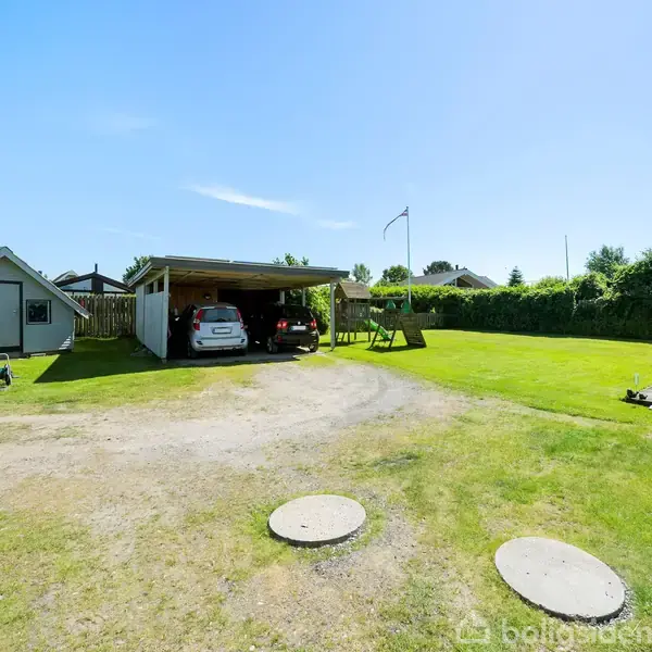 Carport med to parkerede biler, omgivet af en stor grøn græsplæne. Til venstre står et lille hus, og der er en sandet indkørsel. Solskin og blå himmel.