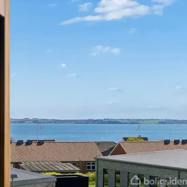 Et stort vindue viser en udsigt over havet. Tagene af flere bygninger ses i forgrunden. Himlen er blå med få skyer.
