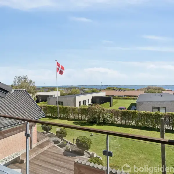Et hus med en altan i forgrunden. På altanen står et glasrækværk; i baggrunden vejer et dansk flag over en have, med flere huse og en blå himmel i horisonten.