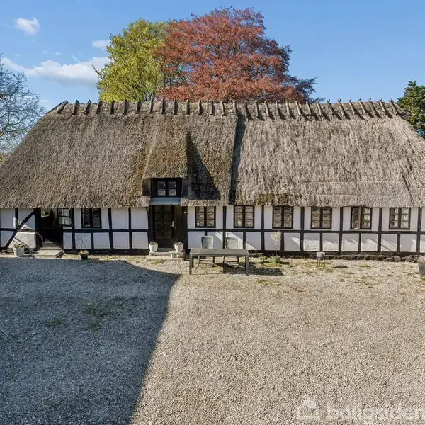 Bindingsværkshus med stråtækt tag står stille på grusplads omgivet af træer. Husets facade har små vinduer og sorte bjælker, der giver et traditionelt, landligt udseende.