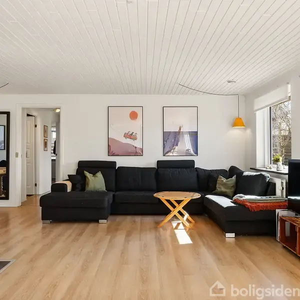 Stor, lys stue med sort L-formet sofa og kakkelbord. To billeder hænger på væggen over sofaen. Der er trælaminatgulv og verdenskort på højre væg ved et stort vindue.