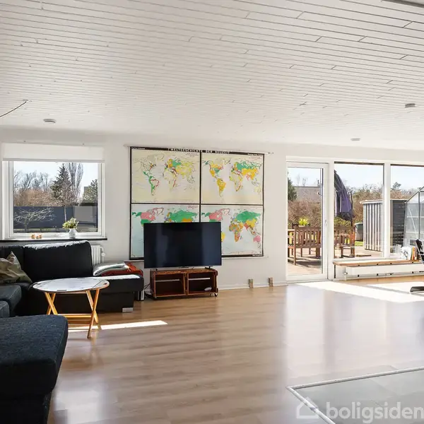 Stue med mørk sofa, bord og TV foran store verdenskort langs væggen. Store vinduer giver udsigt til en terrasse med et væksthus. Lys træparketgulv og hvidt loft.