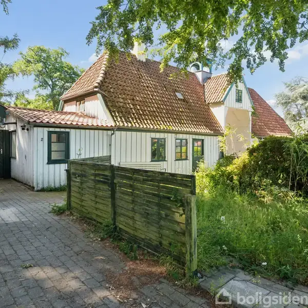 Hus med hvidmalet facade og rødt tegltag står ved siden af en flisebelagt sti, omringet af frodig vegetation. En lille grøn træport markerer grænsen til haven.