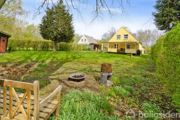 En trægynge står i en have med græs og en bålplads. To huse ses i baggrunden omgivet af træer og buske under en klar, blå himmel.
