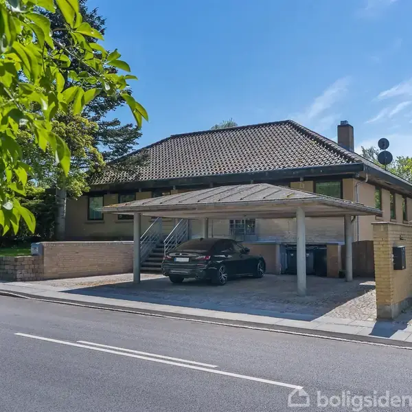 Hus med garage, indeholdende en bil, står ved en vej omkranset af træer og buske under en klar blå himmel.