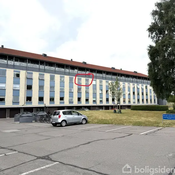 Et boligkompleks med flere etager har en facade med gule og grå striber. En rød cirkel markerer et specifikt vindue. I forgrunden ses en parkeringsplads med en sølvfarvet bil.