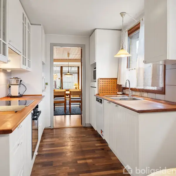 Et køkken med træbordplader og hvide skabe. Rummet er lyst op af naturligt lys gennem vinduer. En åbning fører til en spisestue med træstole og bord.