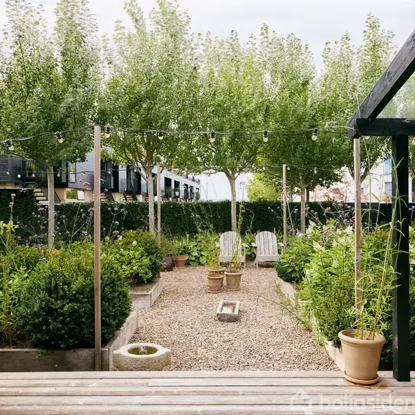 Have: En grussti omgivet af grønne planter og blomster. Stolen er placeret i baggrunden på terrassen af træ. Dekorative lyskæder hænger mellem stolper.
