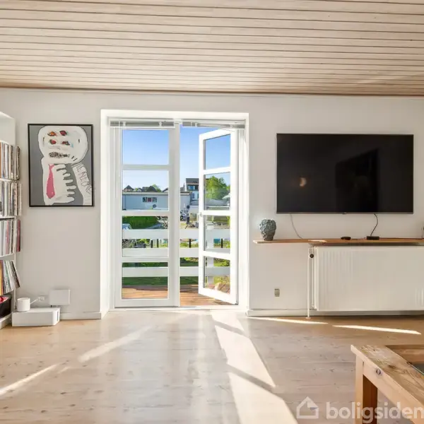 En lys stue med åbne døre til en altan. En bogreol står til venstre, og et stort TV hænger på væggen ved siden af en radiator.