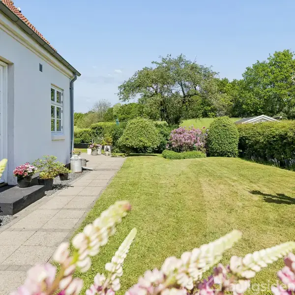 Et hvidt hus ved siden af en flisebelagt sti. Haven er velplejet med grøn græsplæne og buske. Blomster ses i forgrunden, mens træer er i baggrunden.