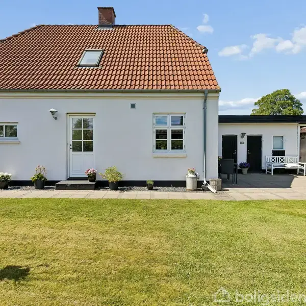 Et hvidt hus med rødt tegltag står stille omgivet af en grøn have og flisebelagt terrasse. Blomsterpotter dekorerer området under et klart blåt himmel.