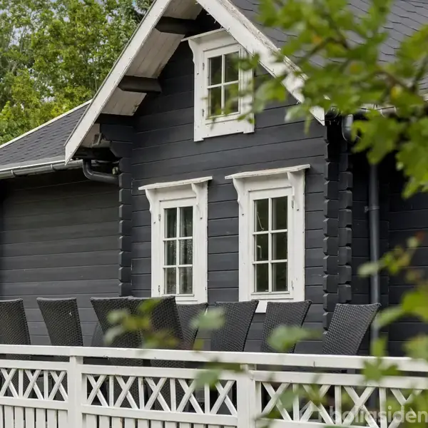 Sort træhus med hvidrammede vinduer og tag, omgivet af en terrasse med sorte stole og hvidt gelænder. Træer og buske i baggrunden.