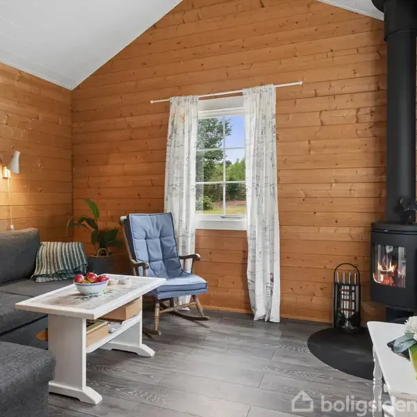 Stue med brændeovn tændt ved siden af blå lænestol og grå sofa. Vindue med hvide gardiner slipper lys ind. Trævægge og gråt gulv skaber hyggelig atmosfære. Bord med blomster.