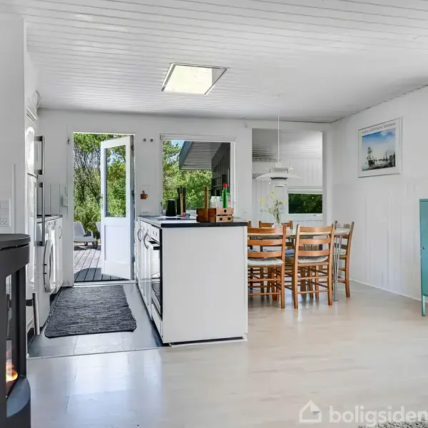 En lys stue med brændeovn til venstre, et spisebord med stole i midten, og en åben dør til en terrasse med grøn udsigt.