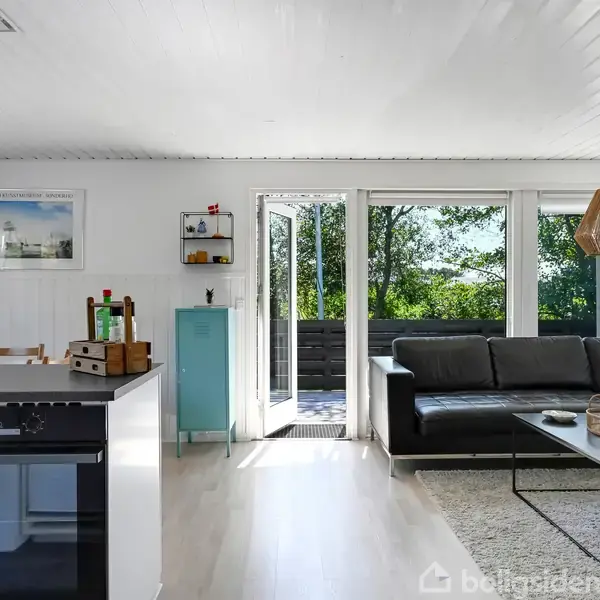 Stue i moderne design. En sofa står mod et vindue med udsigt til en frodig have. Køkkenet har en ø og hængende lampe. Lyst indendørs rum.