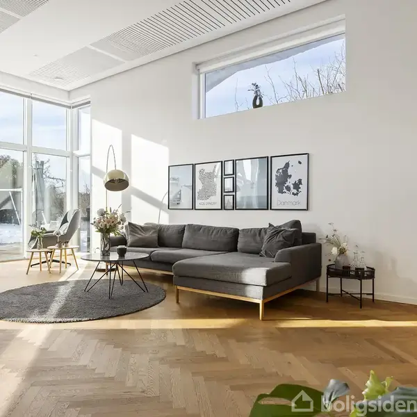 En grå sofa står i en moderne og lys stue med trægulv. Store vinduer giver udsigt til en sneklædt have. Væggen har fem billeder, og rummet er dekoreret med planter.