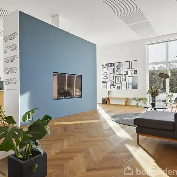 En stue med en væg, der har en indbygget pejs, dominerer rummet. Stuen indeholder moderne møbler, planter og store vinduer med udsigt. Teksten på væggen er ikke synlig i detalje.