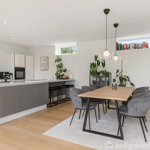 Et moderne køkken-alrum med et stort spisebord omgivet af grå stole. Køkkenø i baggrunden med barstole. Planter og høje vinduer tilfører lys og grønt til rummet.