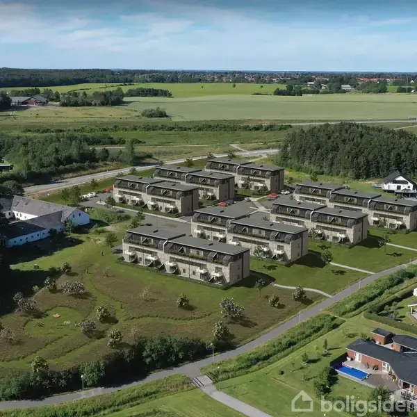 Boligkompleks består af flere rækker moderne bygninger, omgivet af græsarealer og skov. Beliggende i et åbent landligt område med veje og marker i baggrunden.