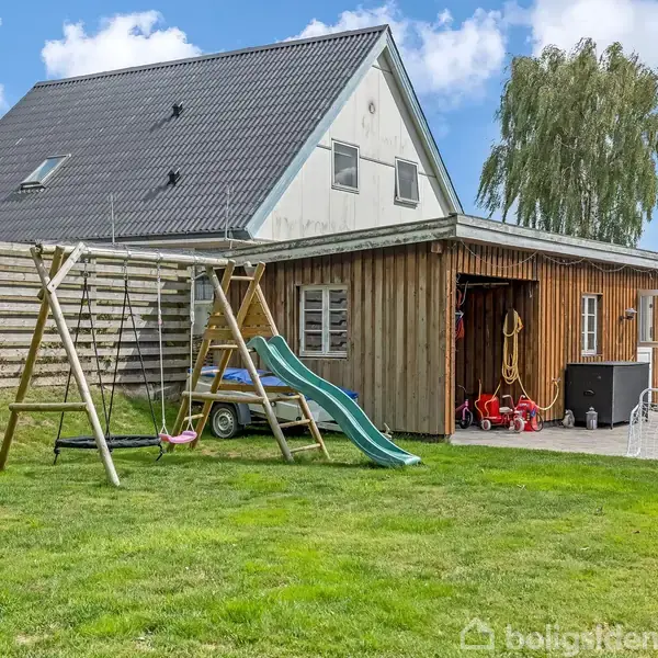 Gyngestativ med rutsjebane står på en grøn græsplæne foran en træbygning. Bygningen har åbne døre med legetøj og haveredskaber. Huset i baggrunden har sort tag.