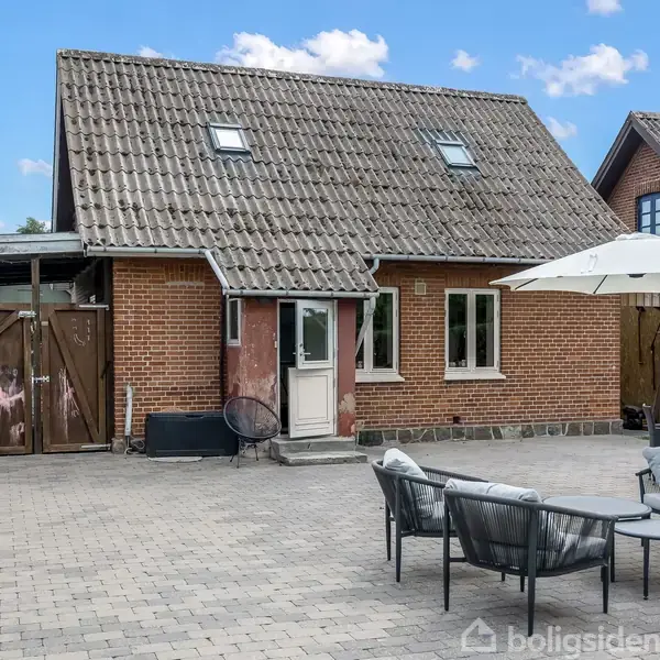 En rød murstensbygning med sadeltag og flere vinduer. Foran huset er en flisebelagt gårdsplads med havemøbler og en parasol. Skyfri himmel i baggrunden.
