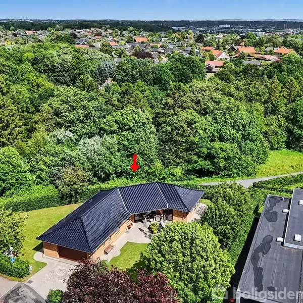 En blå-sort tagbeklædt bolig ligger midt blandt grønne træer. En lille sti løber ved siden af ejendommen. Boligen ligger i et roligt område med tæt vegetation.