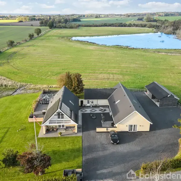 Et stort landhus med gråt tag ligger ved en indkørsel omgivet af græsarealer og træer. I baggrunden ses marker og en stor sø under en skyet himmel.