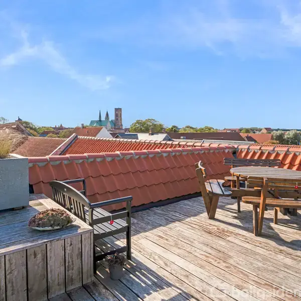 Tagterrasse med træmøbler står på en trægulv, omgivet af røde tegltage. I baggrunden ses kirketårne og en klar blå himmel.