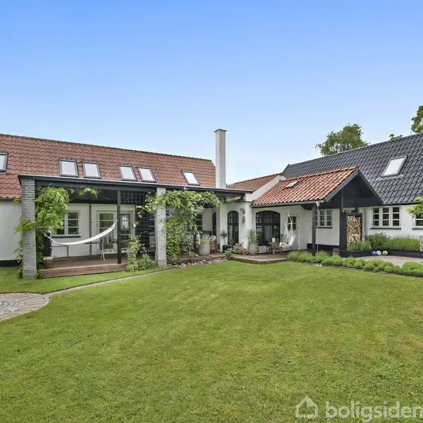 Et lyst hus med rødt og sort tag står stille i en grøn have. Omkring huset er der løvbevoksning og klar blå himmel.