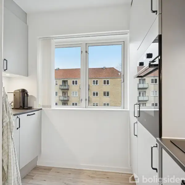 Et stilrent køkken med hvide skabe og sort bordplade. En kaffemaskine står på bordet. I baggrunden ses et vindue med udsigt til boligbyggeri.