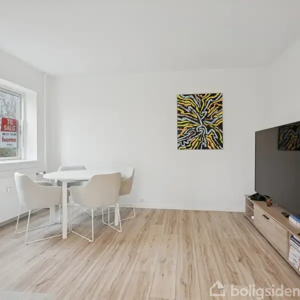 Et moderne, lyst stueområde har et rundt bord med fire stole. Der hænger et farverigt kunstværk på væggen. Et stort TV står på et lavt skab. I vinduet ses teksten: "Til salg home".