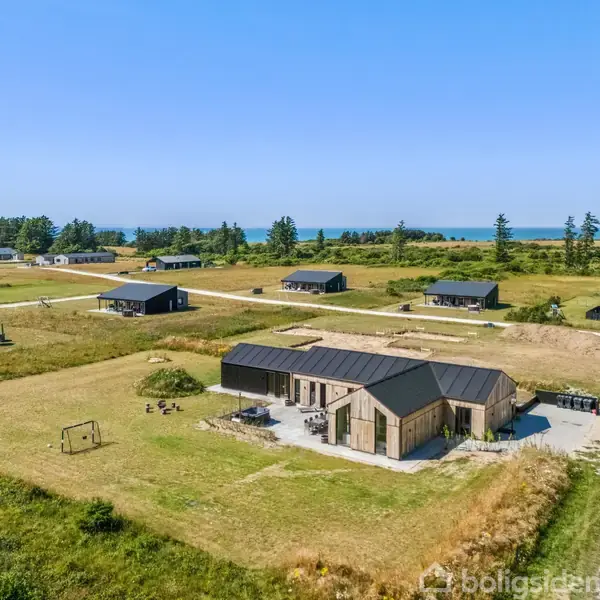 Et moderne hus står på en åben græsplæne med legeredskaber. Huset er omgivet af lignende boliger spredt i et naturskønt område nær skov og hav i baggrunden.